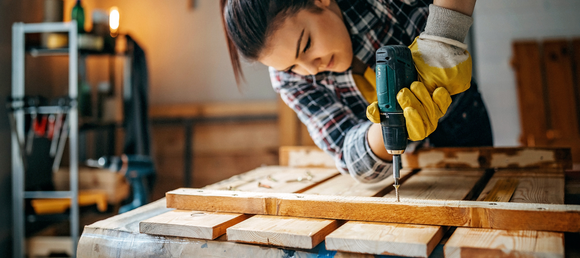 A woman using a drill on her woodworking project in her workshop.